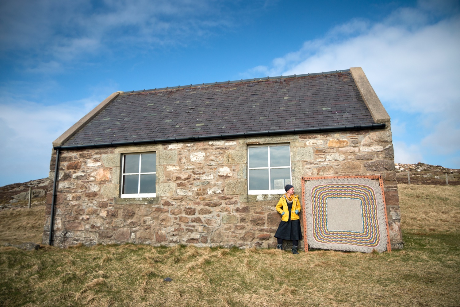 Knitwear designer Kate Davies standing in front of a small stone house with one of her designs
