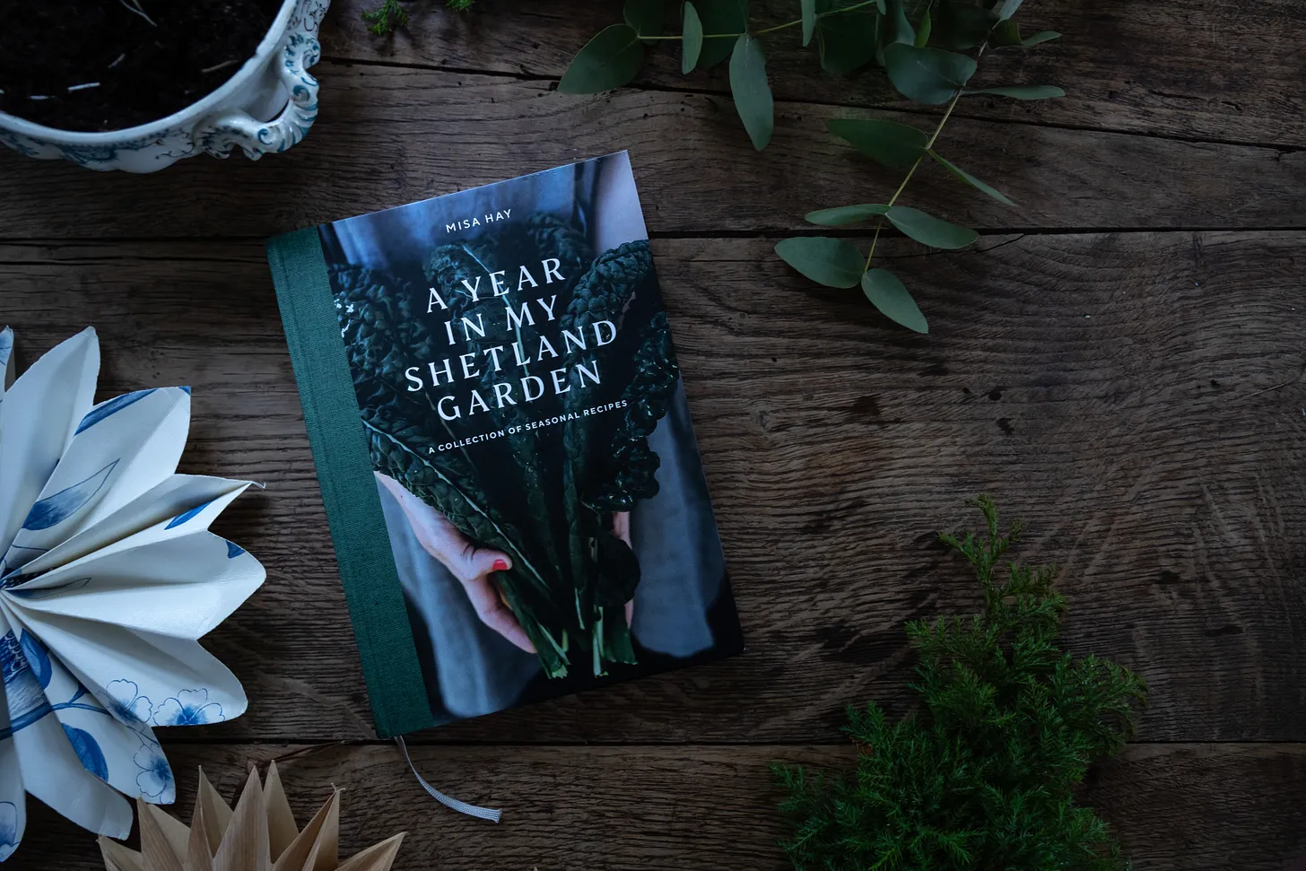 A cookbook lying on a wooden table with Christmas decorations and some natural foliage.