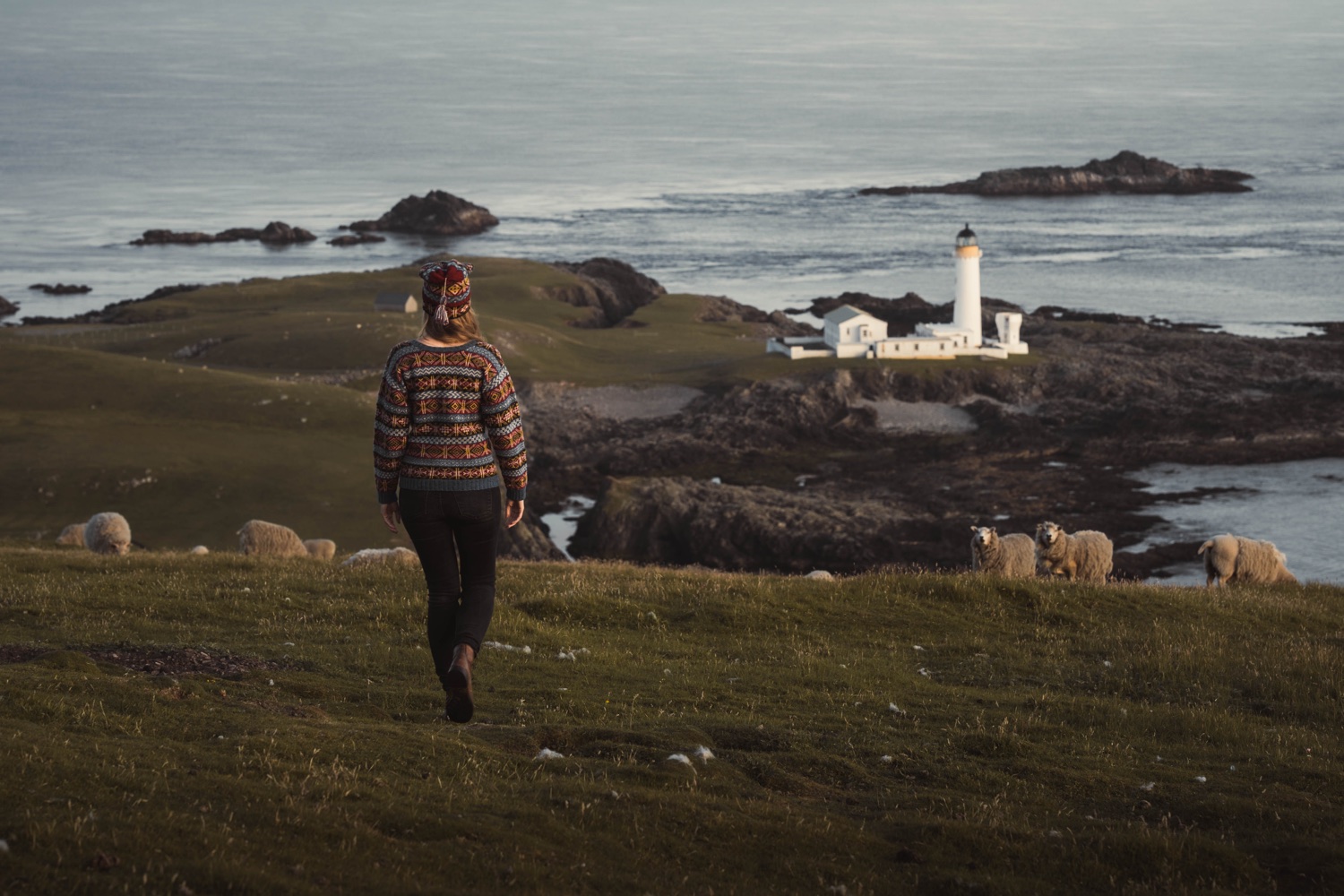 A woman walking towards a lighthouse, wearing a blue and burnt orange Fair Isle jumper. The ocean and sheep surround her.